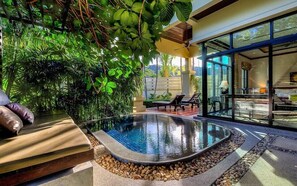 Un espace piscine intérieur moderne, doté d’un jacuzzi, entouré d’une végétation luxuriante et comprenant un coin repas à l’abri derrière des parois de verre.