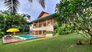 Une maison à deux étages avec une piscine, entourée d’une végétation luxuriante et de palmiers.