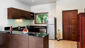 Uma cozinha moderna com armários de madeira escura, bancada preta, micro-ondas branco, geladeira de aço inoxidável e janela com folhagem verde.