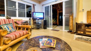 A spacious living room with a large flat-screen TV, a wooden sofa with patterned cushions, a round coffee table with magazines, and a sliding glass door leading to a balcony.