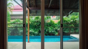 A view through a glass door of a pool area with a tiled floor, surrounded by lush green plants and a thatched roof structure.