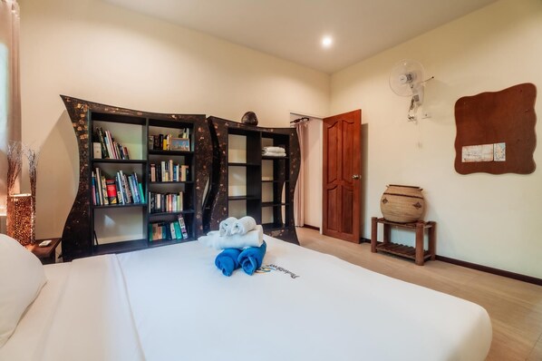 A hotel room with a bed, a bookshelf, a wooden door, a small table with a vase, and a wall-mounted fan.