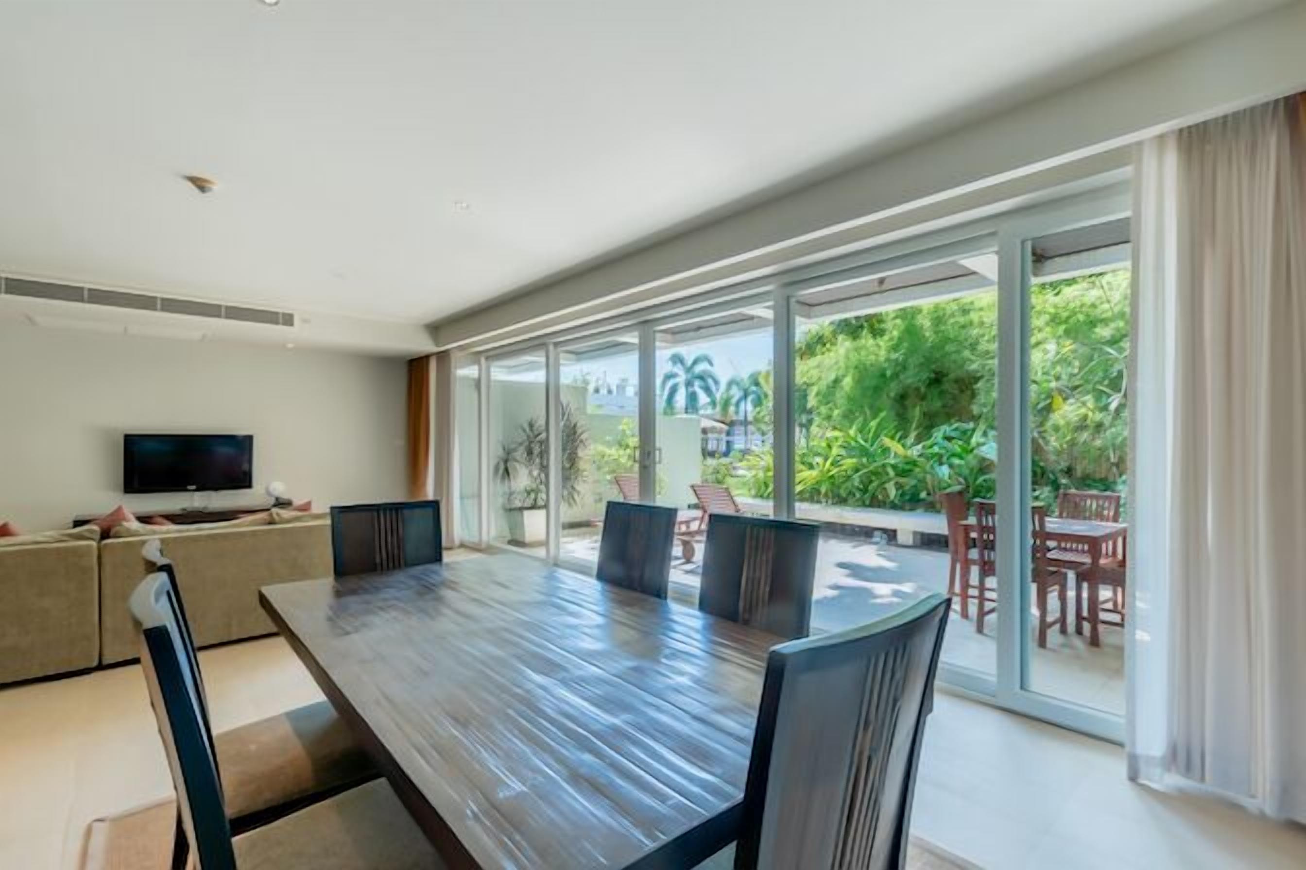 Ein moderner Essbereich mit einem langen Tisch und Stühlen, einem an der Wand befestigten Fernseher und großen Schiebegläsern, die auf eine begrünte Terrasse mit Möbeln führen.