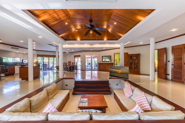 A spacious living area with a high ceiling, a central staircase, and a kitchen in the background.