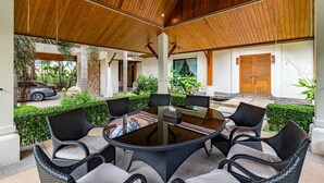 Uma área de refeições ao ar livre com uma mesa redonda rodeada por cadeiras de vime, teto de madeira e vista para o verde e arquitetura circundantes.