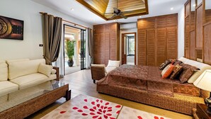 Um quarto espaçoso com uma cama grande, um sofá e uma área de refeições. Há um ventilador de teto, painéis de madeira e vista para o exterior.