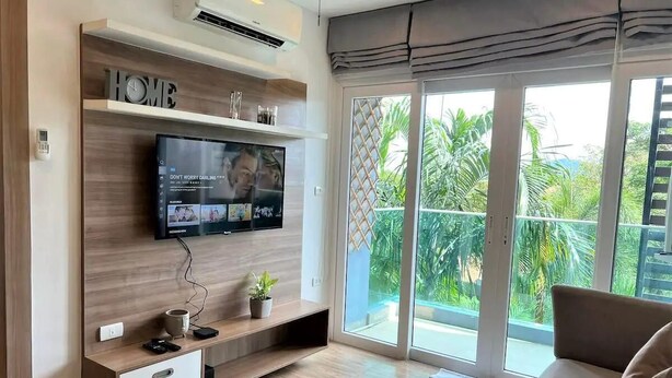 Ett modernt vardagsrum med en platt-TV som är monterad på en vägg av trä, en skjutdörr i glas som leder ut till en balkong med utsikt över grönområden och en beige soffa.