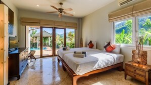 Une chambre spacieuse avec un grand lit, un ventilateur de plafond et une vue sur un jardin luxuriant par la fenêtre.
