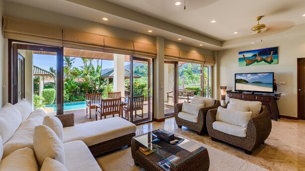 Un salon spacieux comprenant un grand canapé, deux fauteuils, une table basse et un téléviseur. La pièce est dotée de grandes portes-fenêtres coulissantes qui donnent sur un espace piscine aménagé avec du mobilier d’extérieur.