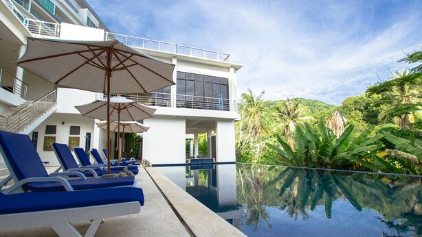 A poolside area with lounge chairs and umbrellas, a pool reflecting the surrounding greenery, and a building with balconies and large windows.