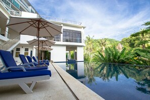A poolside area with lounge chairs and umbrellas, a pool reflecting the surrounding greenery, and a building with balconies and large windows.