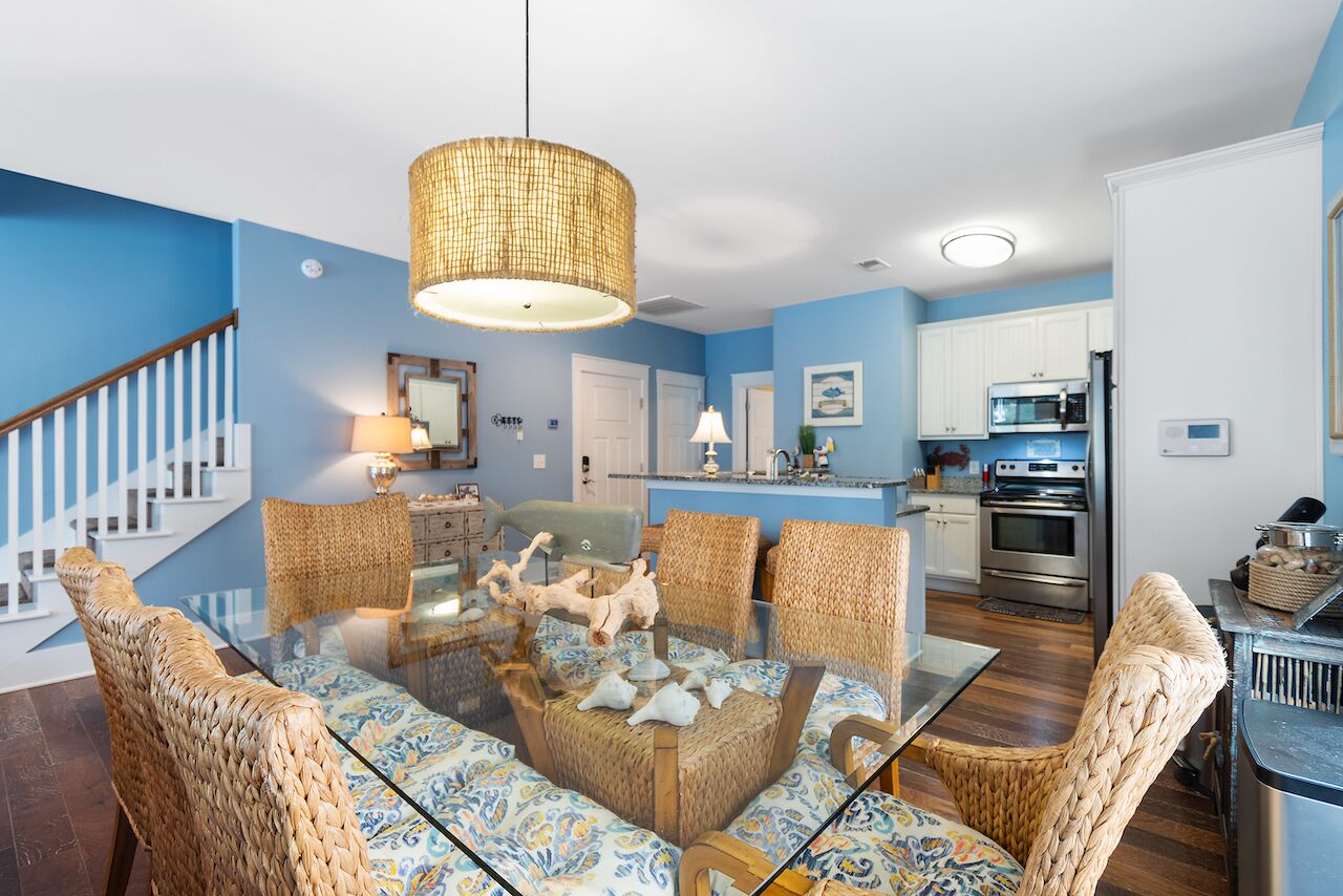 A dining area with a glass table surrounded by wicker chairs, a hanging light fixture, and a kitchen in the background.