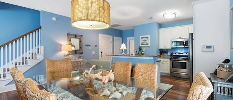 A dining area with a glass table surrounded by wicker chairs, a hanging light fixture, and a kitchen in the background.