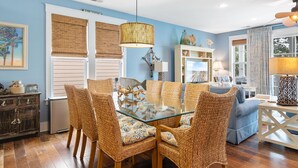 Un comedor con mesa de vidrio rodeada de sillas de mimbre, lámpara colgante y espejo decorativo sobre la mesa.
