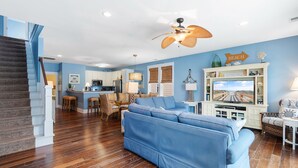 Un salón espacioso con sofá azul, suelo de madera, una cocina al fondo y una escalera a la izquierda.