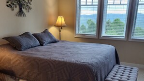 A bedroom with a large bed, a nightstand, a lamp, and a view of the outdoors through three windows.