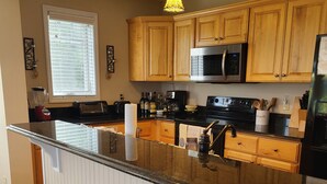 A kitchen with wooden cabinets, a black countertop, a stainless steel microwave, and a bar stool.