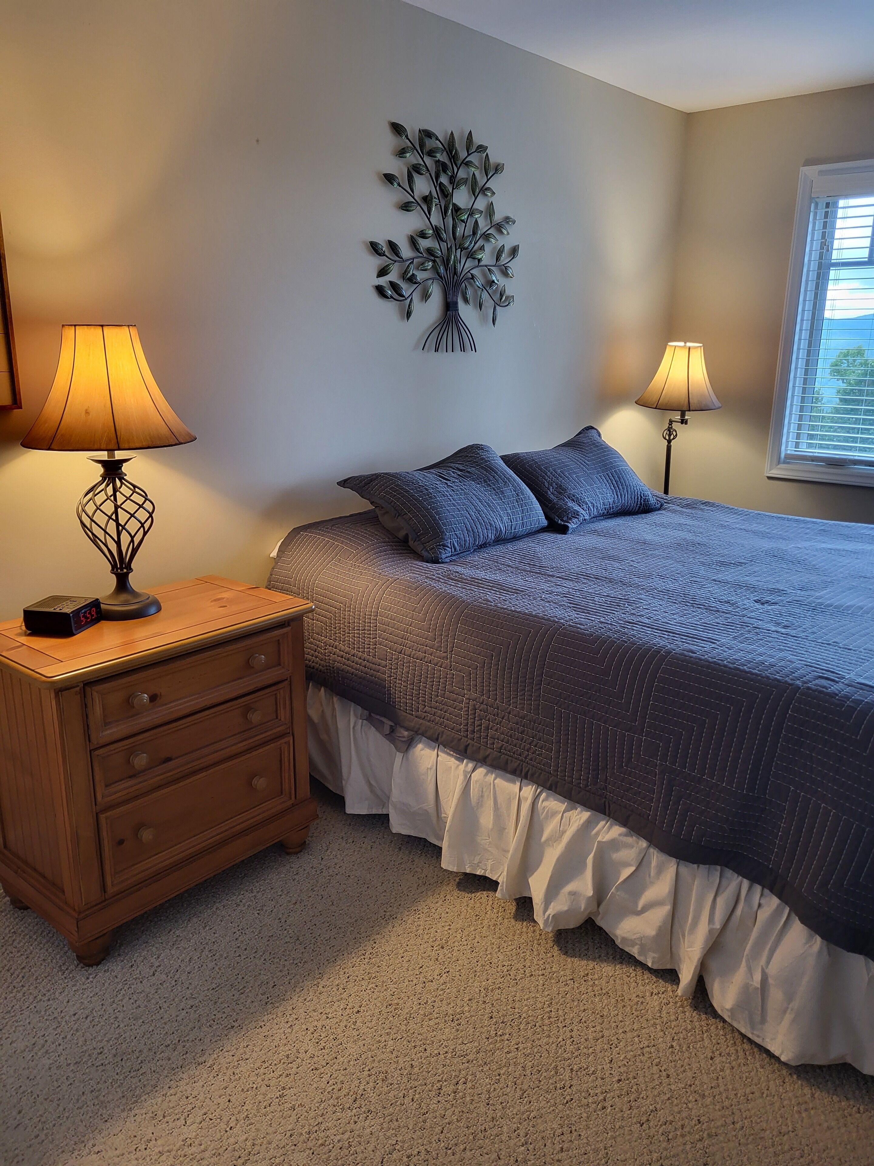 A bedroom with a bed, two bedside tables, a wall clock, a window, and a wall decoration.
