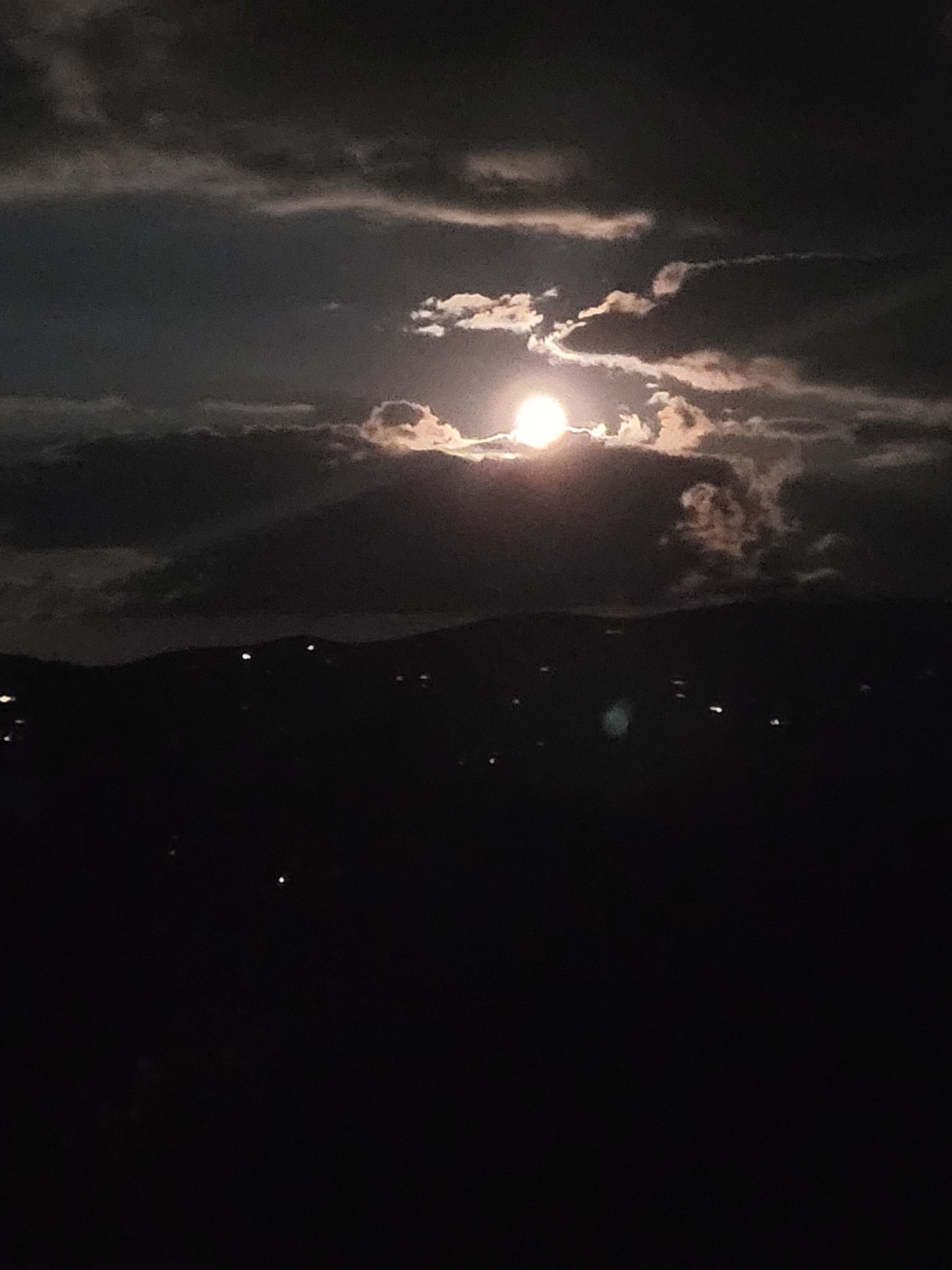 A night sky with a bright moon partially obscured by clouds, and a silhouette of a mountain range below.