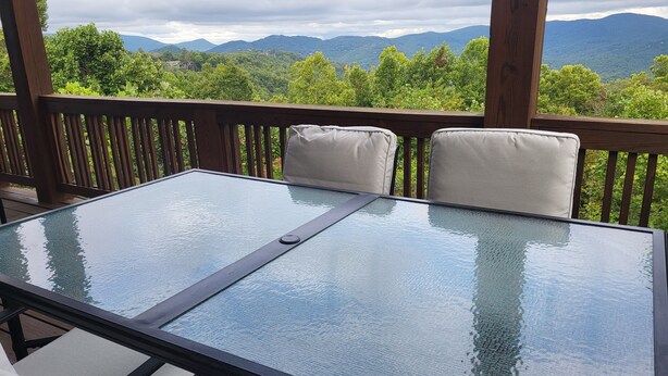 A glass-top table with two cushioned chairs on a wooden deck, overlooking a mountainous landscape.