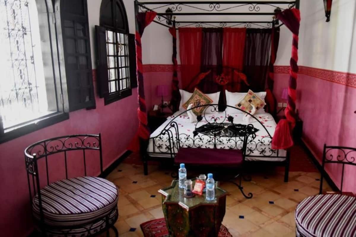 A room with a four-poster bed, two chairs, a small table with water bottles, and a window with decorative shutters.