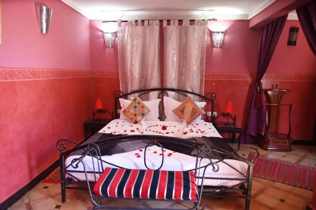 A bed with a metal frame, white bedding, and decorative pillows. There are two bedside tables with lamps. The room has pink walls and a patterned rug.