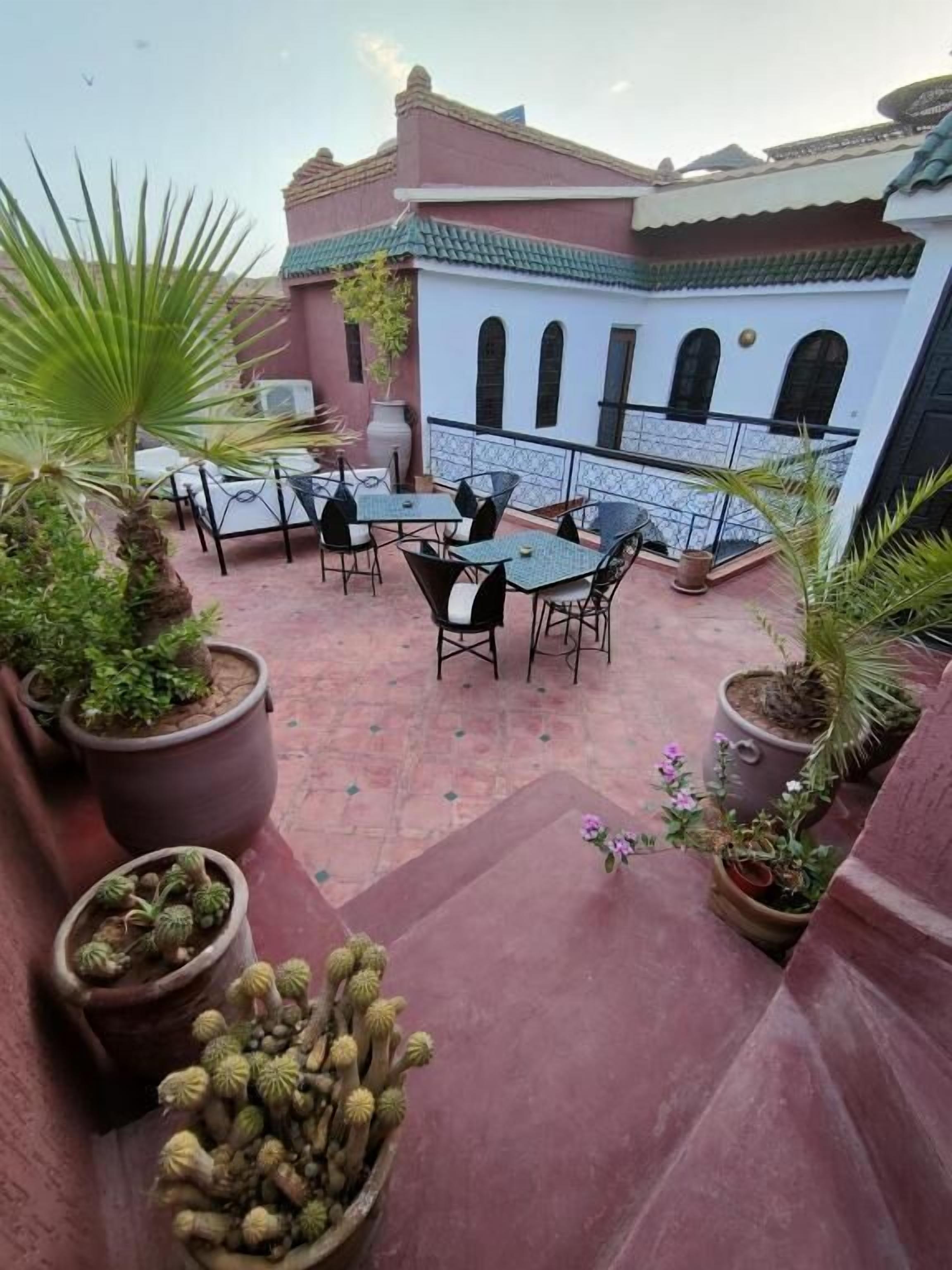 Un espace extérieur aménagé en terrasse, avec plusieurs tables et chaises, entouré de plantes en pot et, au fond, d’un bâtiment dont le toit est recouvert de tuiles.