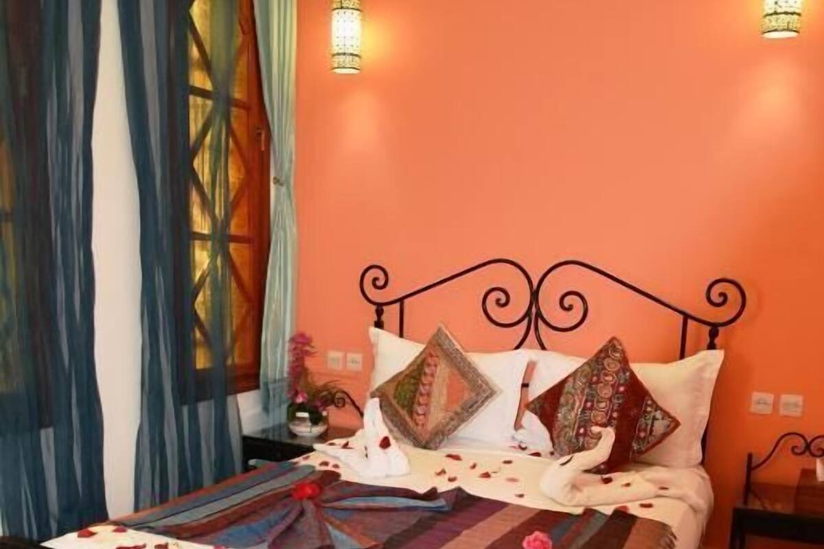 A bed with a patterned bedspread, pillows, and a decorative item on the side. There is a small table with water bottles and a glass.