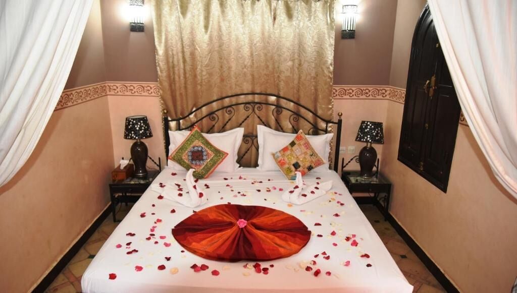 A bed with rose petals and a red decorative pillow, two wall-mounted lamps, and a curtain.