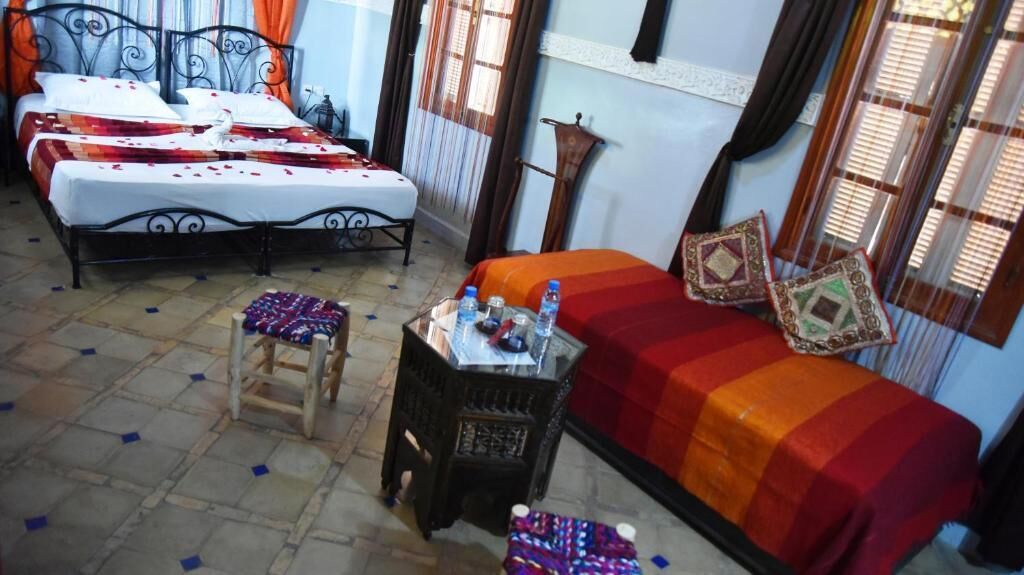 A hotel room with two beds, a small table with a chair, and a window with curtains.