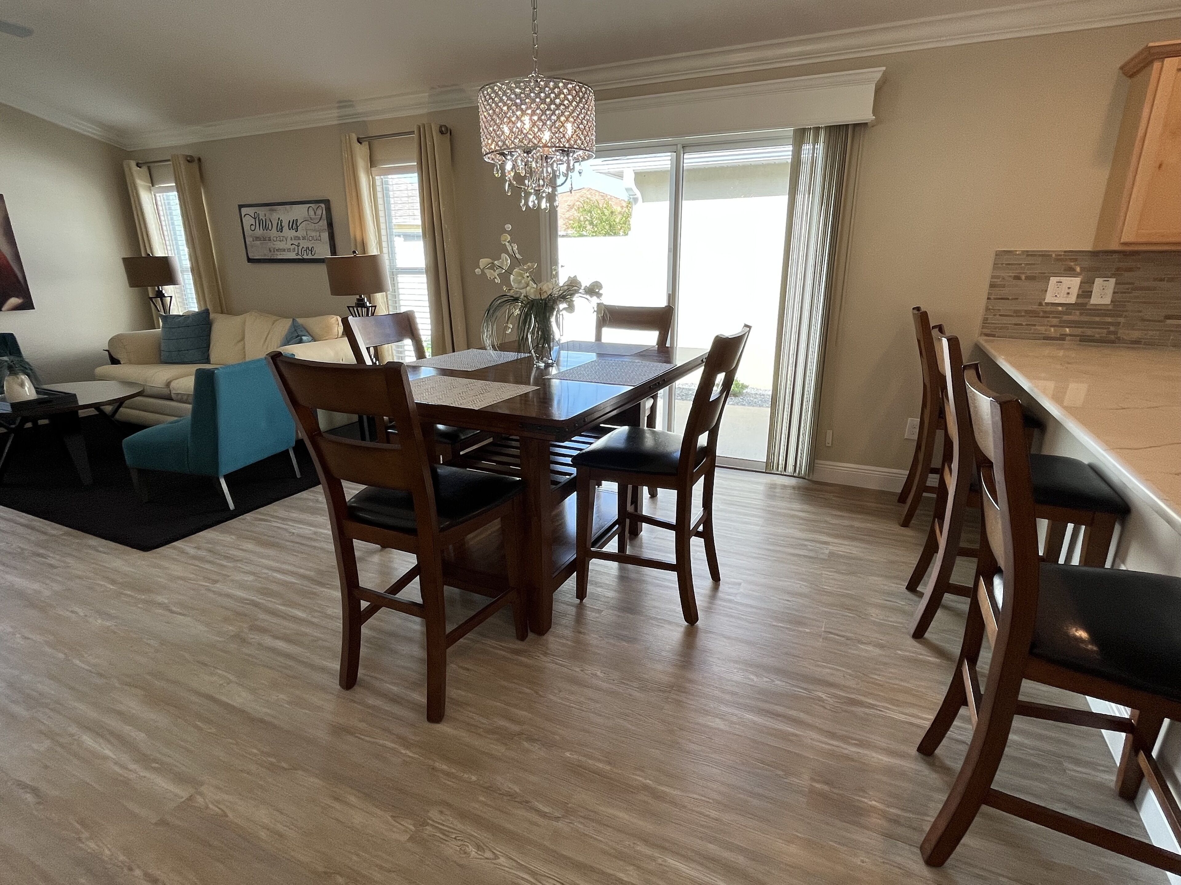 A dining area with a wooden table and chairs, a chandelier, and a view of the outdoors through large windows.