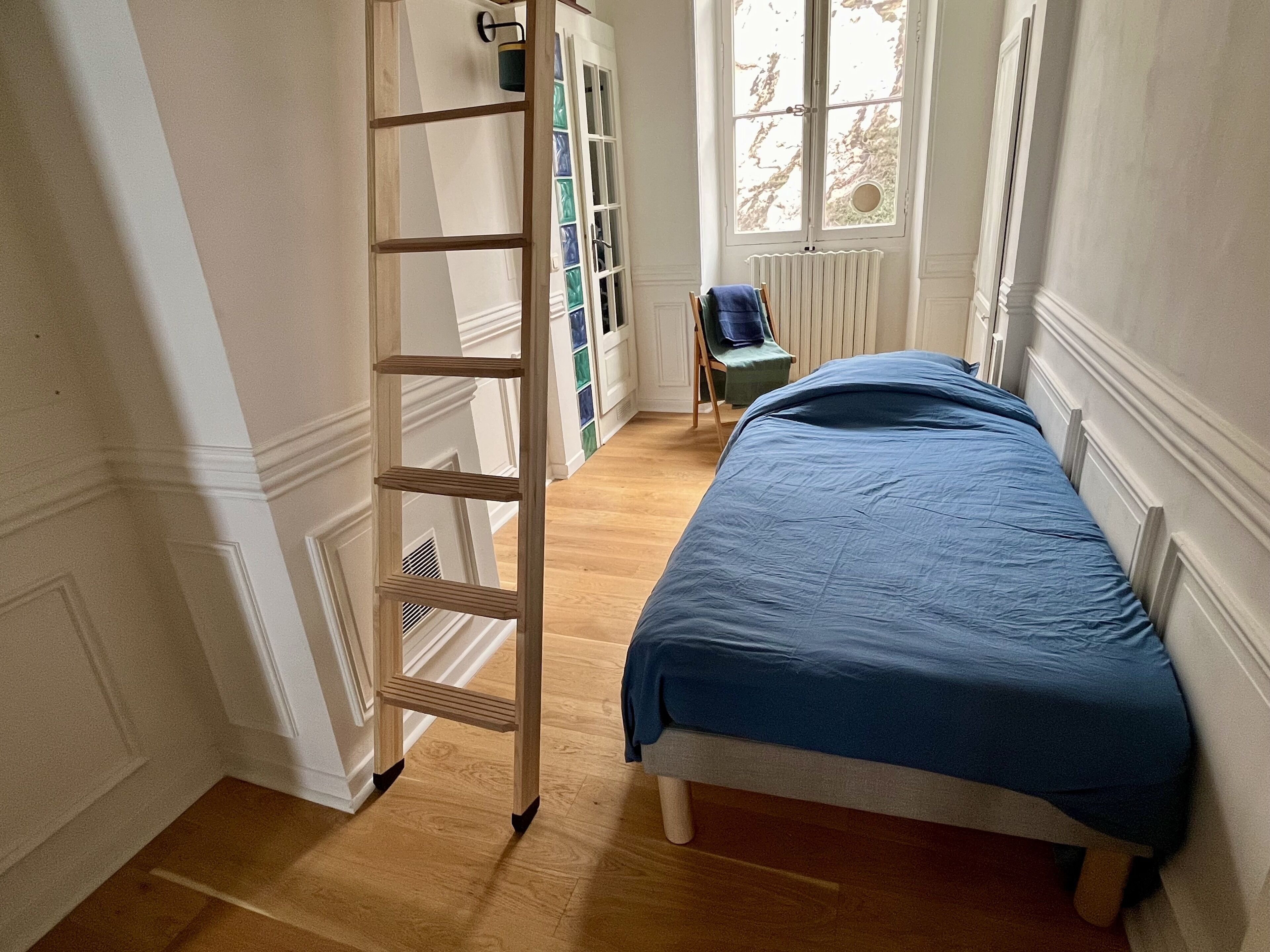 A bedroom with a wooden ladder, a bed with a blue blanket, a radiator, and a window with a view of trees.