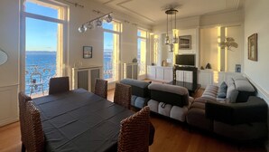 Ett rymligt vardagsrum med ett stort matbord, bekväma sittplatser och utsikt över havet genom öppna fönster.