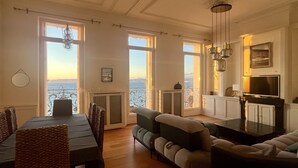 Ett rymligt vardagsrum med ett matbord, soffor och en TV. Stora fönster ger utsikt över havet.