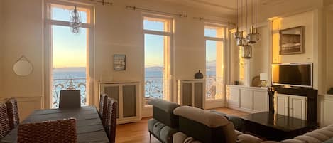 A spacious living area with a dining table, sofas, and a TV. Large windows offer views of the sea.