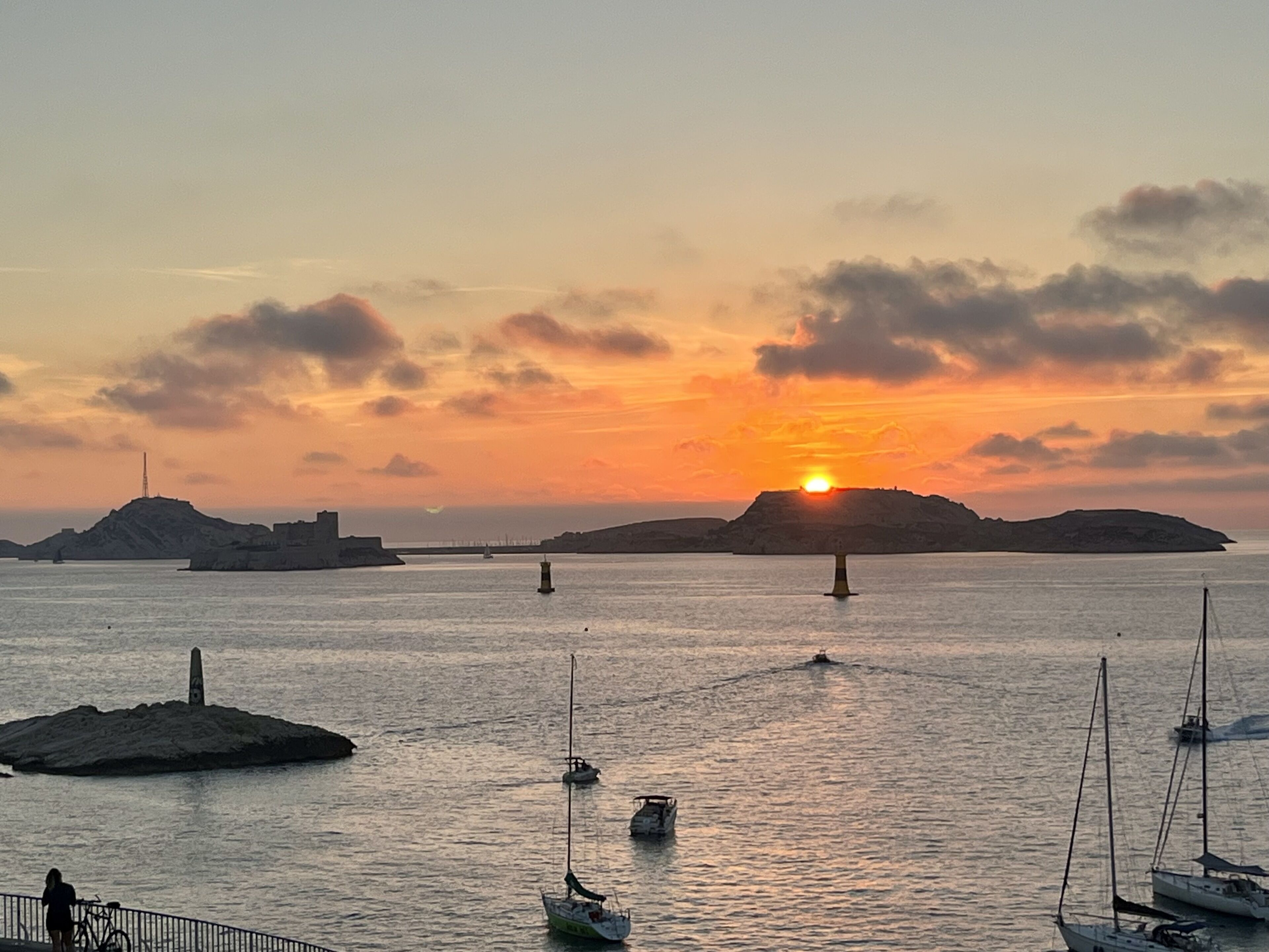 Un coucher de soleil sur une mer calme, avec plusieurs bateaux et un phare au loin.