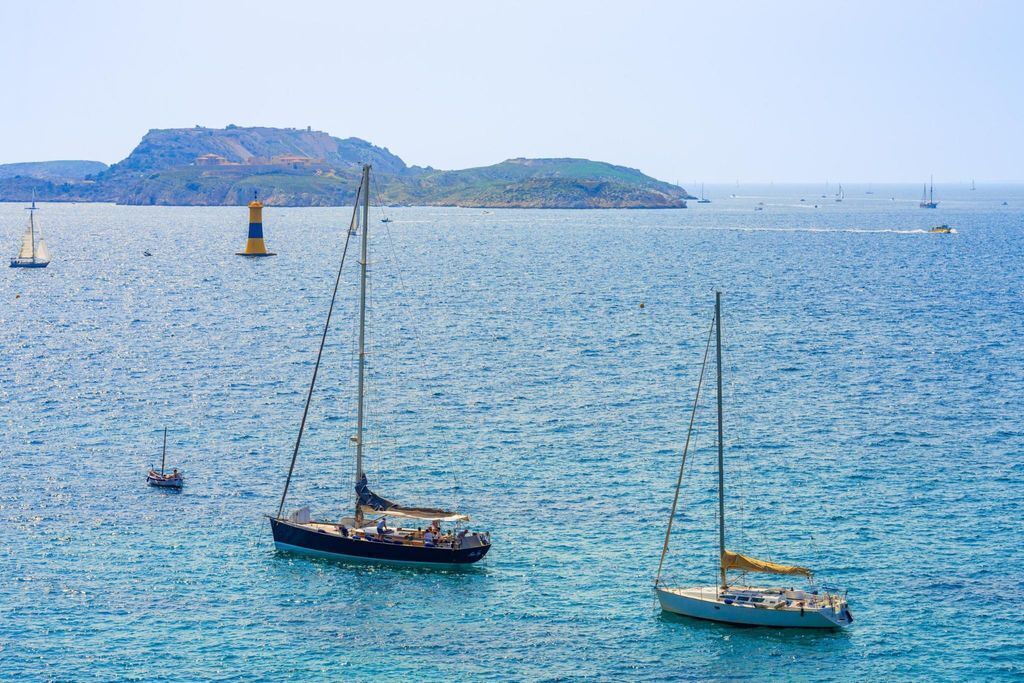 Un paysage côtier avec des voiliers sur une eau calme, un phare et une île rocheuse en arrière-plan.