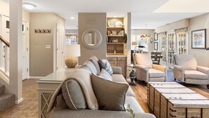 A spacious living room with a large sectional sofa, a coffee table, and a fireplace. There is a kitchen area visible in the background with a dining table and chairs.