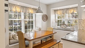 A dining area with a wooden table and chairs, large windows with blinds, and a hanging pendant light.