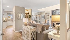 A spacious living area with a sofa, a glass coffee table, and a fireplace. There is a kitchen in the background with white cabinets and a dining area with a table and chairs.