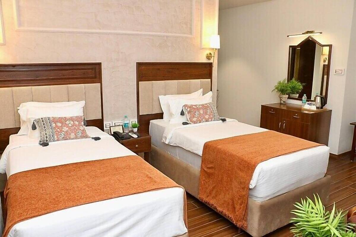 Une chambre d’hôtel avec deux lits, chacun doté d’une tête de lit en bois, de draps blancs et d’un plaid marron. On y trouve une table de chevet avec une lampe, ainsi qu’une petite table sur laquelle sont posés une plante et un miroir.