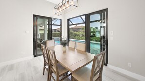 Un espace repas avec une table et des chaises en bois, un luminaire suspendu et de grandes portes-fenêtres coulissantes donnant sur une piscine.