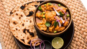 Un tazón de curry con coliflor, decorado con cilantro y rodajas de cebolla roja, acompañado de pan naan y un gajo de lima sobre un tapete tejido.