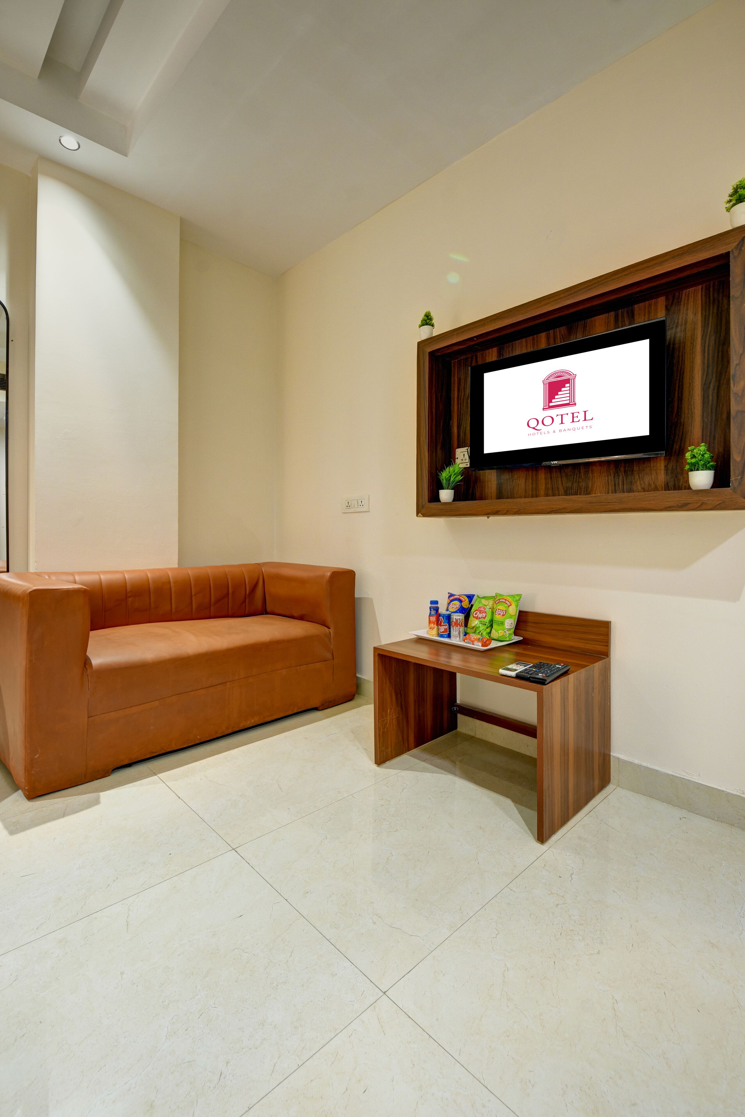 A living room with a brown leather sofa, a wooden coffee table with snacks, a television on the wall, and a small potted plant.