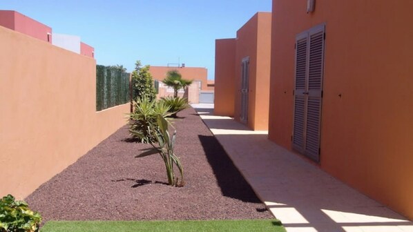 A narrow alleyway with a gravel path, bordered by buildings with orange walls and shutters, and a small palm tree in the center. - Villa Sabine con Piscina, Vista Campo Golf y mar (Antigua)