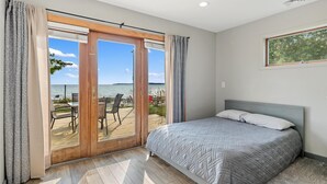 Un dormitorio con cama, vista al mar a través de un ventanal corredero y una pequeña mesa con sillas afuera.