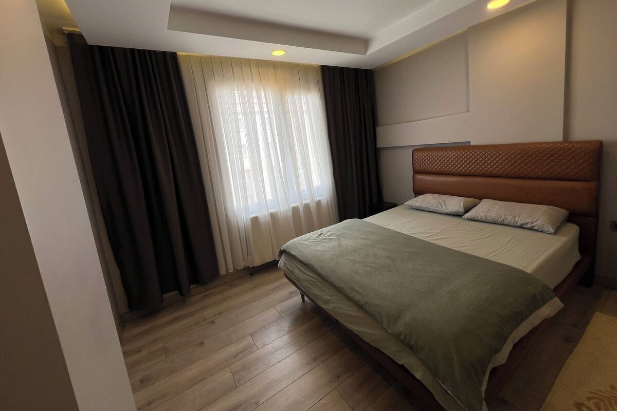Ett modernt hotellrum med trägolv, en säng med ett vadderat sänggavel och ett stort fönster med gardiner.
