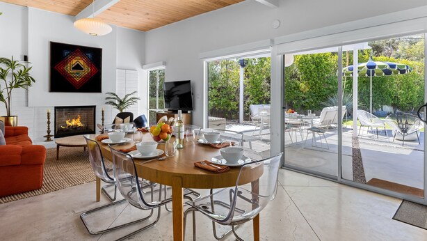 A modern dining area with a wooden table, clear chairs, a fireplace, and a view of the outdoors. - Racquet Club Rendezvous – Palm Springs Escape (Palm Springs)