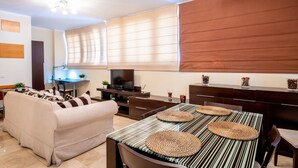 A modern living room with a sofa, a dining table with woven placemats, a TV, and a kitchen area with a wooden cabinet. - Céntrico Apartamento en Santa Cruz (Santa Cruz de Tenerife)