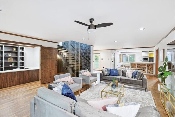A spacious living room with a modern ceiling fan, a staircase with a unique design, and a kitchen in the background.